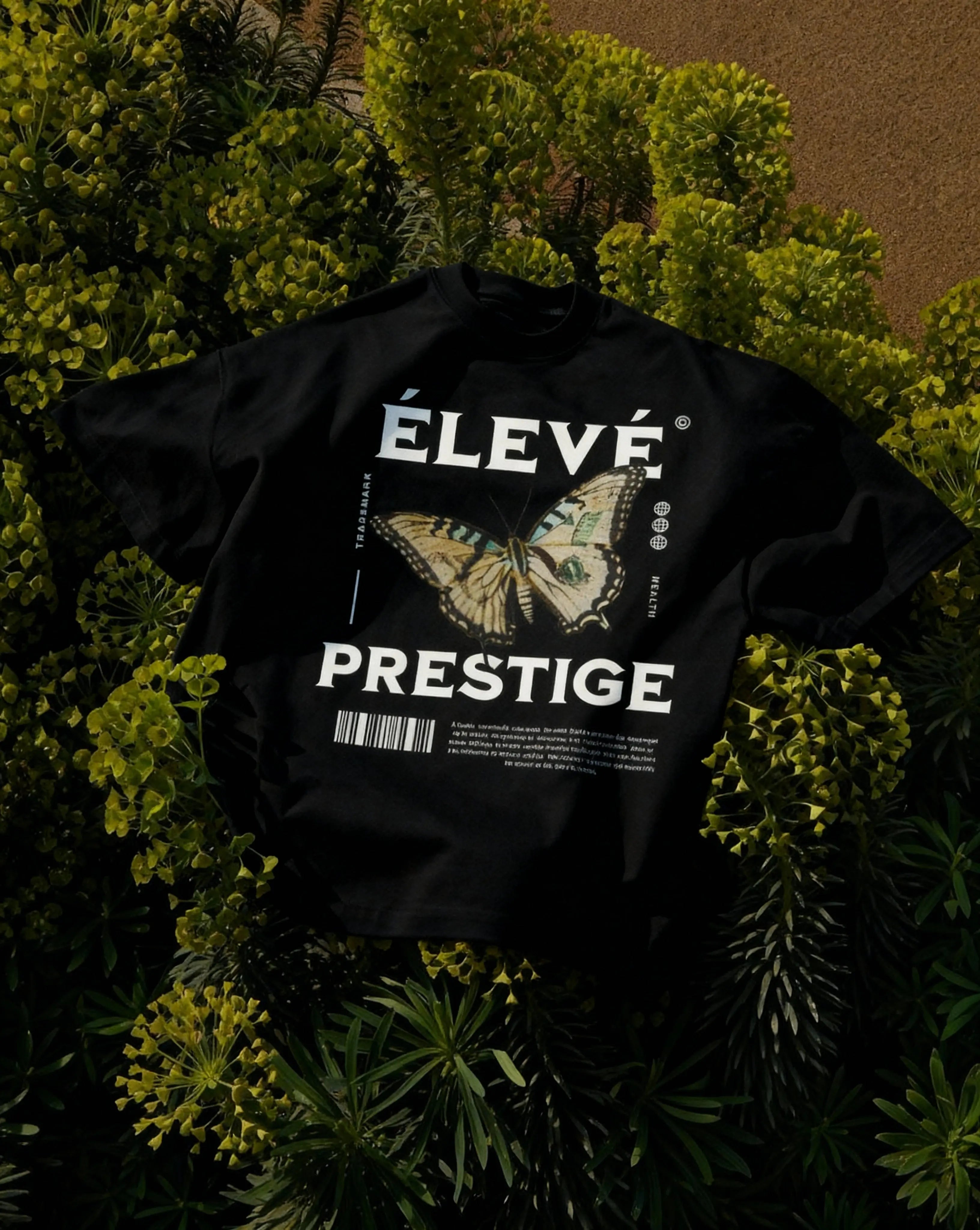 ÉLEVÉ Prestige Butterfly T‑Shirt Black mit großem Butterfly Frontprint, liegend in Pflanzen – Premium Streetwear Produktfoto aus dem Prestige Drop