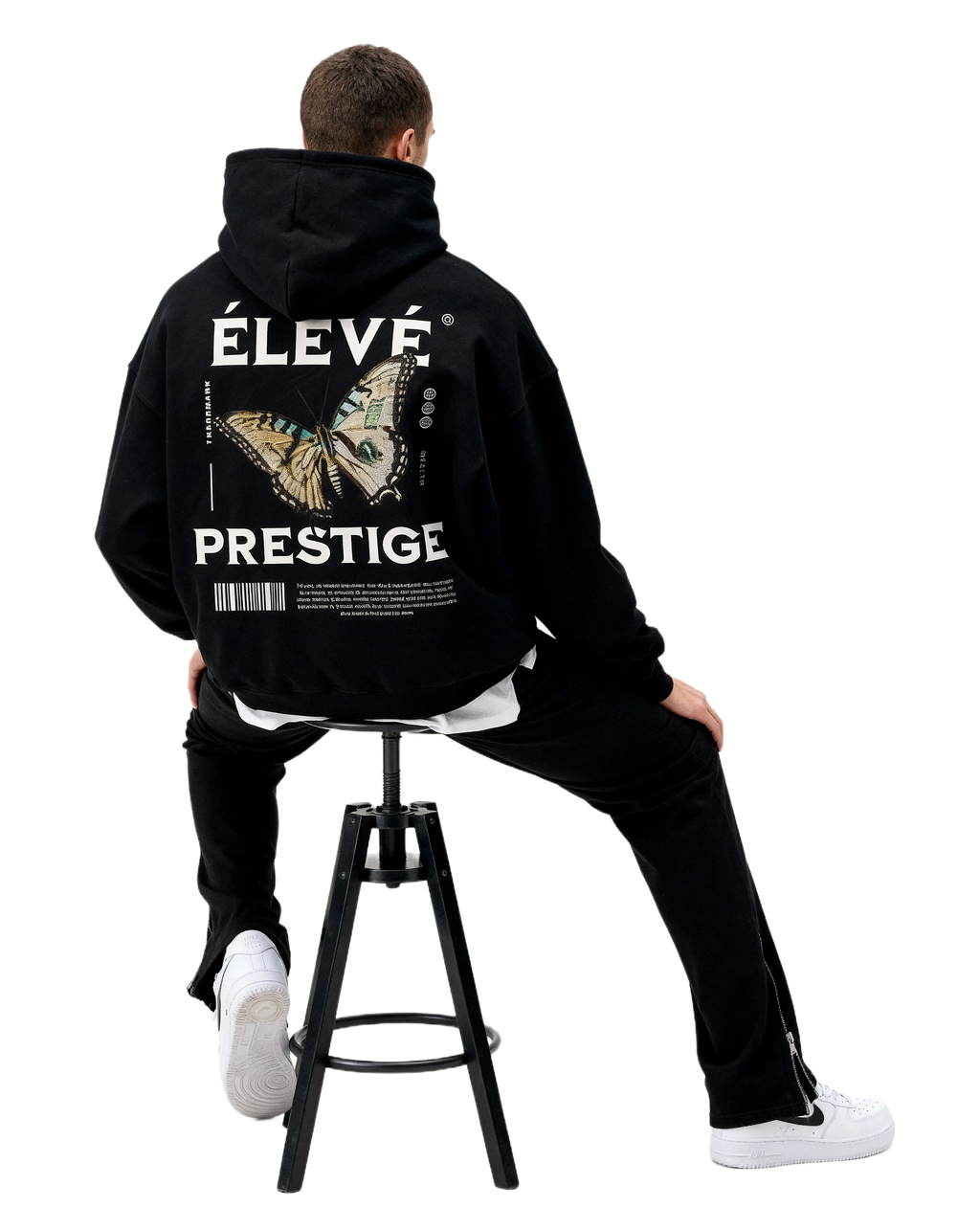 ÉLEVÉ Prestige Hoodie Black mit großem Butterfly Backprint, Model sitzend auf Hocker – schwarzer Premium Unisex Streetwear Hoodie aus dem Prestige Drop